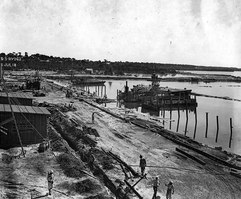 Historical black and white photograph showing construction workers at a waterfront site with a barge and wooden pilings in the background.