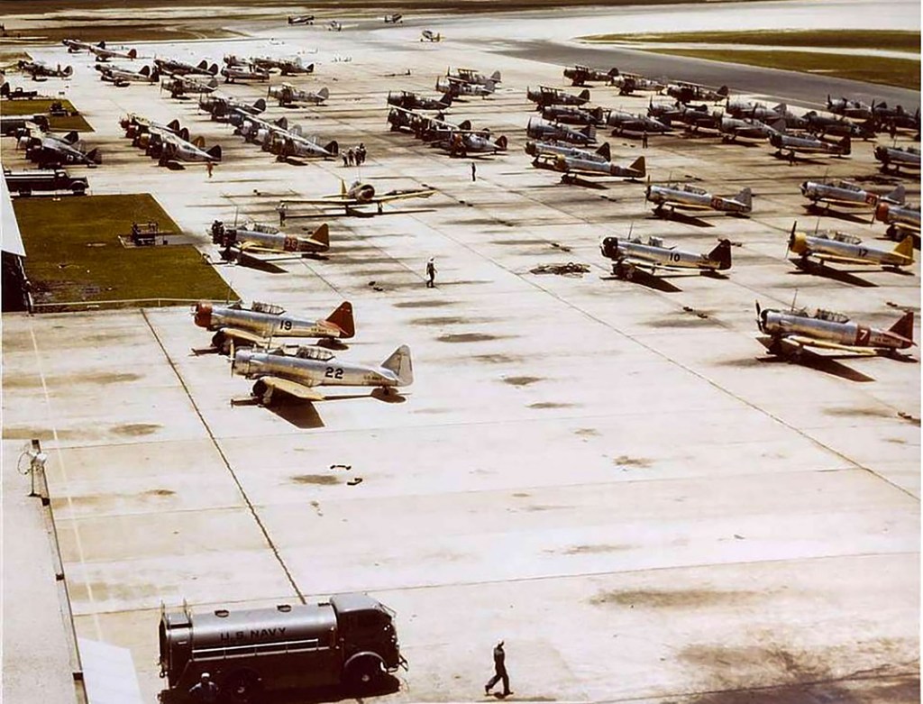 Aerial view of an airfield filled with various vintage aircraft, with personnel working and a military fuel truck parked nearby.