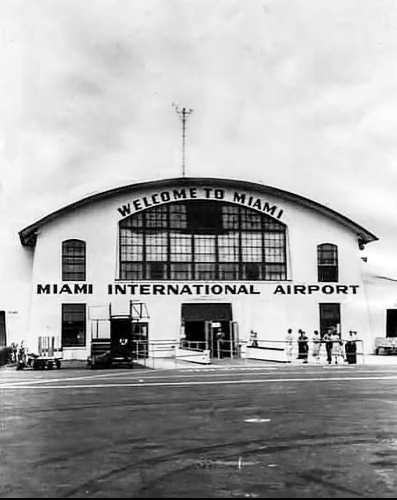 Historic black and white photograph of the entrance to Miami International Airport, featuring a large archway sign that reads 'Welcome to Miami' and 'Miami International Airport'.
