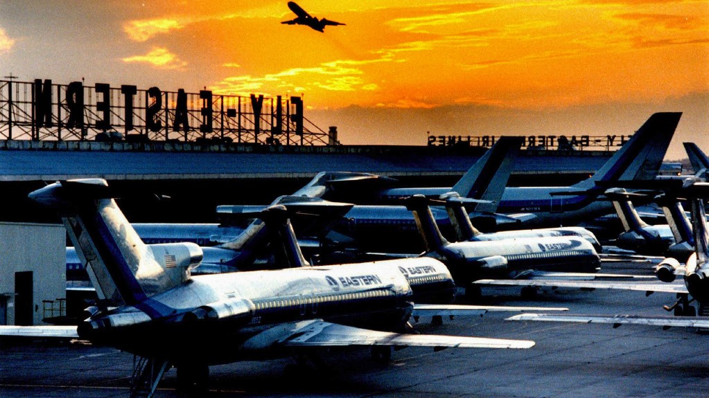Aerial view of an airport during sunset featuring multiple Eastern Airlines planes on the tarmac with an airliner taking off in the background.
