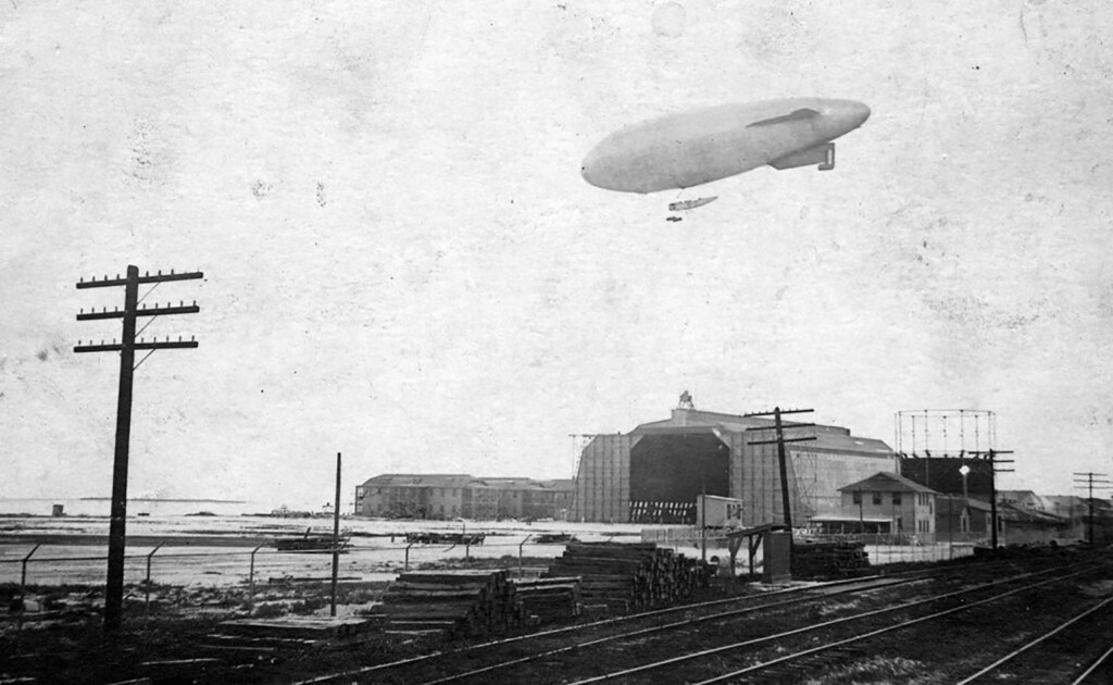 Historic black and white photograph of a zeppelin flying above a hangar with power lines and a railway in the foreground.